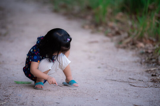 Cute Girl Playing Stick On Ground. Child Draw Pictures On Soil In Middle Of Nature. Concept Allows Kids To Play And Learn With Nature As They Imagine. Asian Children Wear Blue Shirt Is 3-4 Years Old.