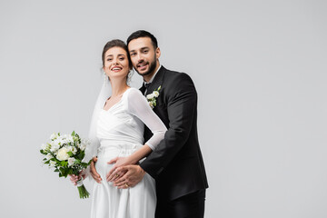 Young muslim groom embracing cheerful pregnant bride with wedding bouquet isolated on grey