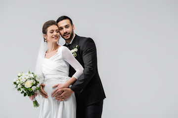 Smiling pregnant bride standing near muslim groom isolated on grey