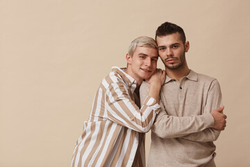 Minimal waist up portrait of happy gay couple looking at camera while posing against beige...
