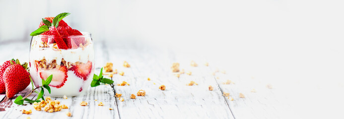 Banner with healthy breakfast of strawberry parfaits made with fresh fruit, yogurt and granola over a rustic white table. Selective focus on glass jar in front with blurred background. 