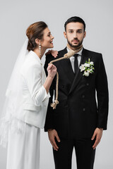 Cheerful bride in wedding clothes holding slip knot on neck of muslim groom isolated on grey