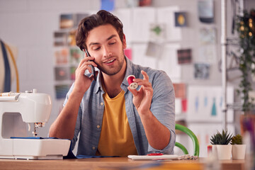 Male Owner Of Fashion Business With Mobile Taking Phone Call At Sewing Machine In Studio