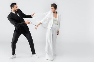 Smiling muslim man in suit pulling veil and handcuffs of bride on white background