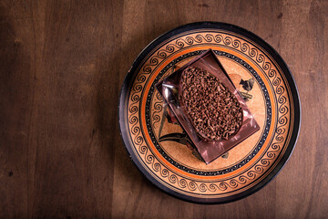 Top view of a stuffed chocolate easter egg with grated chocolate on the top and a spoon on the side in a 
transparent package on a plate with an african design on a wooden table.
