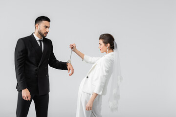 Muslim man in handcuffs looking at bride in veil isolated on grey
