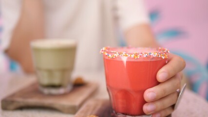 Hand picking up a glass of pink smoothie decorated with sprinkles. Ice coffee served on the wooden tray in the background