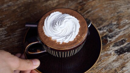Hand serving a brown mug of plant based hot chocolate with whipped cream on a wooden table