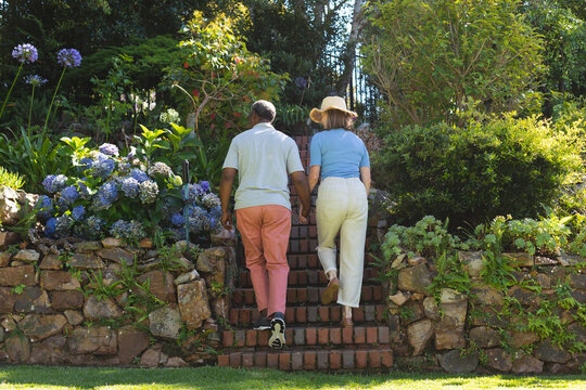 Diverse senior couple walking in garden holding hands