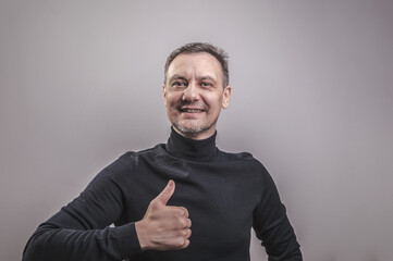 A white man of European appearance middle aged with gray in his hair and a slightly unshaven face in a black turtleneck happily gives a thumbs up
