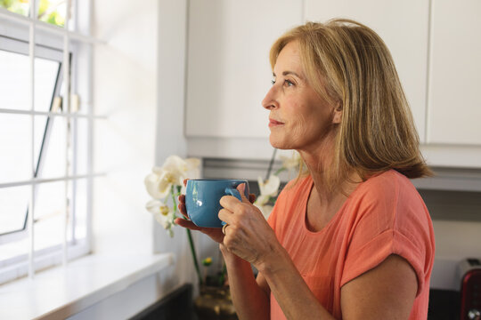 Caucasian senior woman standing by window drinking cup of coffee