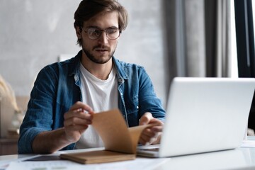Serious young businessman with the glasses sitting and using laptop at home office and taking notes.