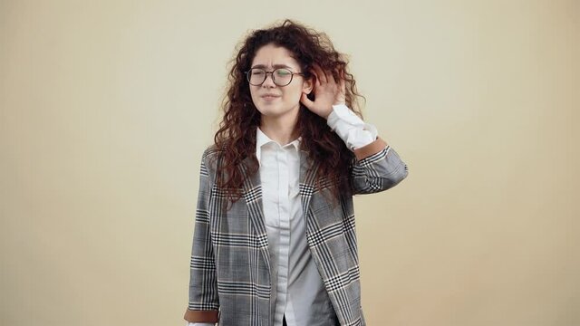 The curious young woman, with her hand to her ear, listens to the soup, unwilling to hear well. Cretaceous in gray jacket and white shirt, with glasses posing isolated on a beige background in the