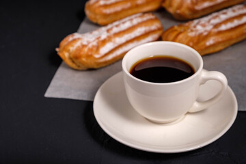 Black coffee in a white cup and eclairs with cream on a dark background
