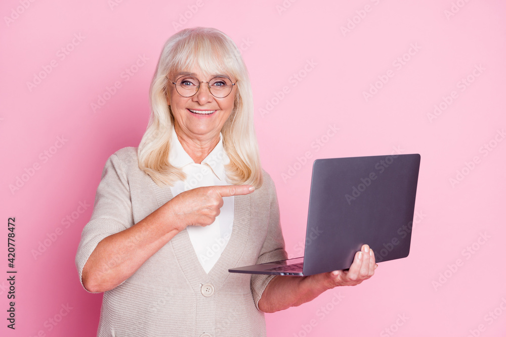 Wall mural Photo of old lady hold computer direct finger wear spectacles beige cardigan isolated pastel pink color background