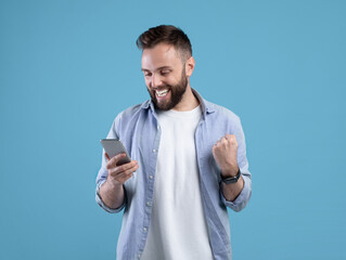 Portrait of happy bearded young man using smartphone, making winner gesture over blue studio background