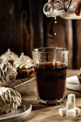 Pouring milk into a glass of hot coffe. In focus the front of a glass and a hand with a bottle.