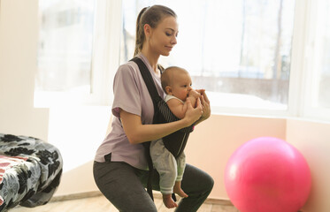 A young mother performs sports exercises squats and lunges with the child on the chest in the baby carrier. The joys and difficulties of motherhood