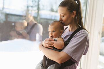 A young mother with a child in her arms stands by the window. Happy family. Motherhood during quarantine. Home insulation