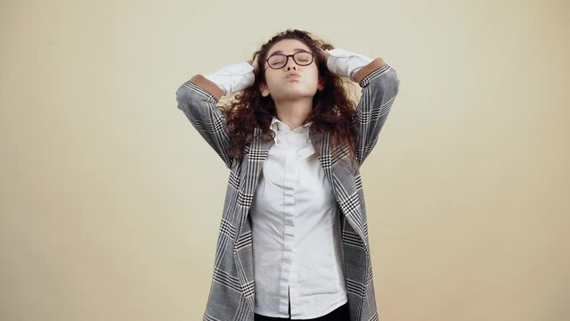 Exhausted young woman, tired with her hands in her hair, breathing hard. Cretaceous in gray jacket and white shirt, with glasses posing isolated on a beige background in the studio. The concept of