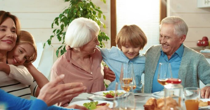 Happy family sitting at dinner table. Little grandson hugs grandparents, daughter hugs mom. Concept of family weekend, holiday table, meeting of generations.