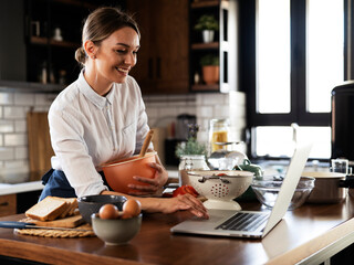 Young woman cooking in the kitchen. Beautiful woman following recipe on laptop and preparing delicious food.