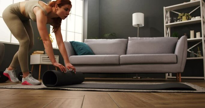 Fit young woman in sports clothes unrolling fitness mat on floor before doing yoga practice. Lady getting ready for home workout.