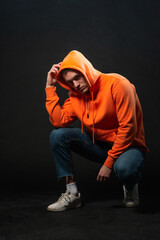 Male model sitting on the floor and posing in orange sports hoodie isolated over a black background.