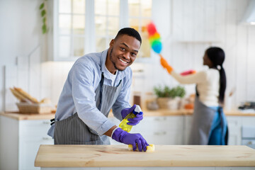 Handsome black guy washing kitchen table while his girlfriend dusting on background, free space