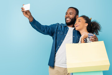 happy african american man taking selfie with cheerful wife holding shopping bags on blue