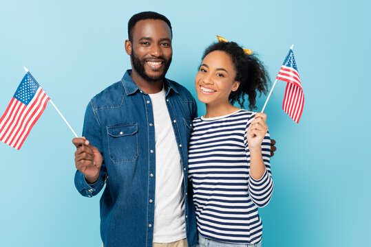 cheerful african american couple holding small flags of usa while looking at camera isolated on blue