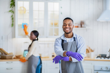Obraz premium Portrait of positive African American guy with mop ready for cleanup, his wife washing kitchen cabinet on background