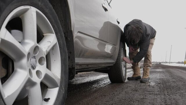 Man on the side of the road changing a tire of his car that broke down during winter. He is removing the wheel nuts