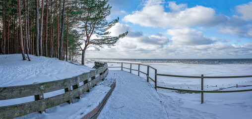 Panoramic view of wooden bench and Baltic sea coast covered in snow during sunny day with blue sky and clouds. Covered in snow sea beach with pine forest in winter.