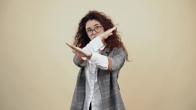 The serious, responsible young woman says stop crossing her hands as a gesture, to stop. Cretaceous in gray jacket and white shirt, with glasses posing isolated on a beige background in the studio
