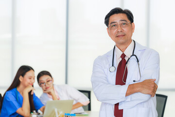 Fototapeta premium Portrait senior male doctor wearing eye glasses with background of young female doctor, and a young nurse with laptop are meeting in a meeting room in the hospital.