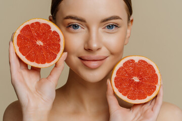 Close up shot of healthy lovely woman holds halves of grapefruit near face has healthy glowing skin...