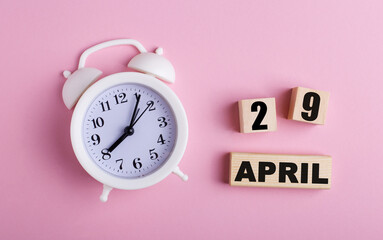 On a pink background, a white alarm clock and wooden cubes with the date of APRIL 29