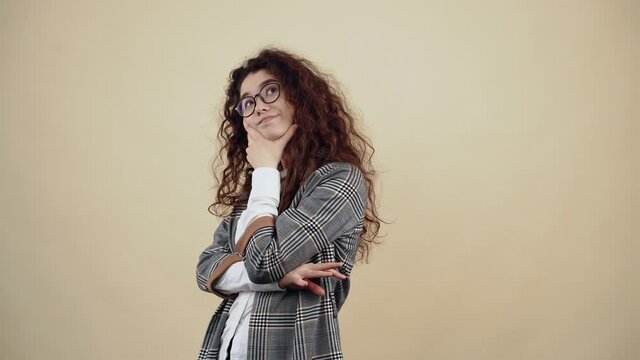 The beautiful young woman, with her arms embraced, thinks of new solutions, at the end she smiles with her teeth. Cretaceous in gray jacket and white shirt, with glasses posing isolated on a beige