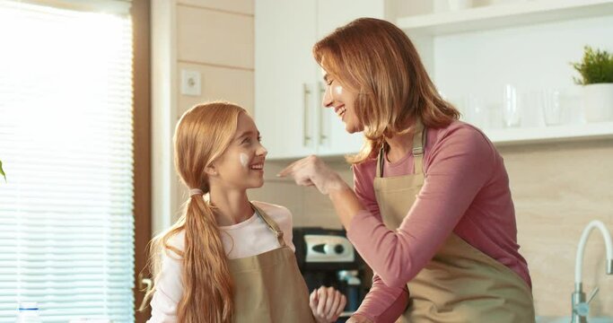 Cheerful mother spends free time in kitchen with her daughter preparing cookies and having fun smearing their faces with flour on each other.