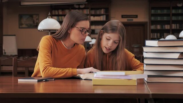 Attractive older sister in a yellow sweater and glasses explain sister lessons the teacher sits in the school library with schoolgirl and helps to teach homework distance learning self isolation.