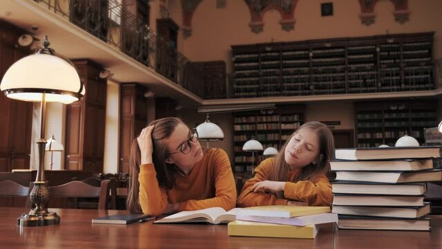 Two charming sisters in identical yellow sweaters fell asleep at a table in a classic school library Share ideas study together communicating cooperating in classroom sit at desk distance learning.