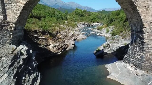 Drone flies through a bridge along the river. Mountains of Corsica and the evergreen scrub forest - macchia can be seen.