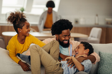 Afro father and children are tickling on the sofa in the living room