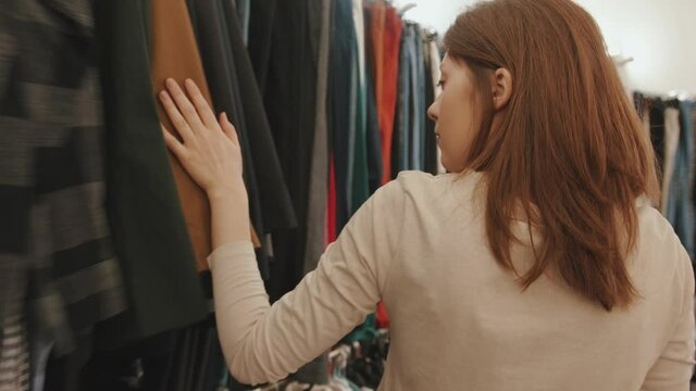 A sensual young girl while shopping in a boutique walks past the rows of clothes and chooses an outfit. Back view