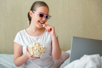 Teenage girl is watching movie on laptop and eating popcorn while sitting in bed. Young happy woman watching movie or TV series at home.