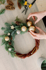 Making rustic christmas wreath, seasonal winter workshop. Florist hands holding pine cone and making christmas wreath on wooden table with festive decorations. Holiday advent