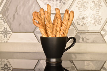 Several grissini breadsticks in a black cup, close-up, on a background of tiles.