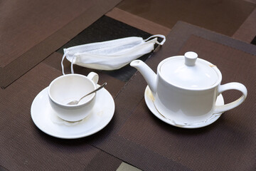 Teapot and a cup on a saucer on dark napkins on the table. Next to the medical protective mask