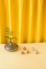 A gray vase with palm leaves, next to a quail egg. Yellow curtain in the background. Easter concept 2021.